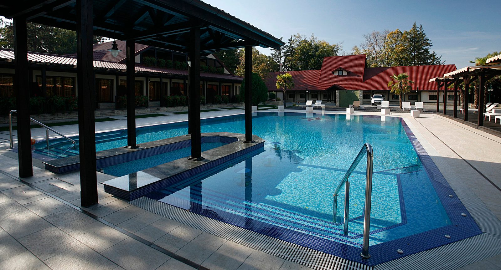 Piscina văzută de sub pergola de lemn, cu bancheta din apă și clubul în fundal
