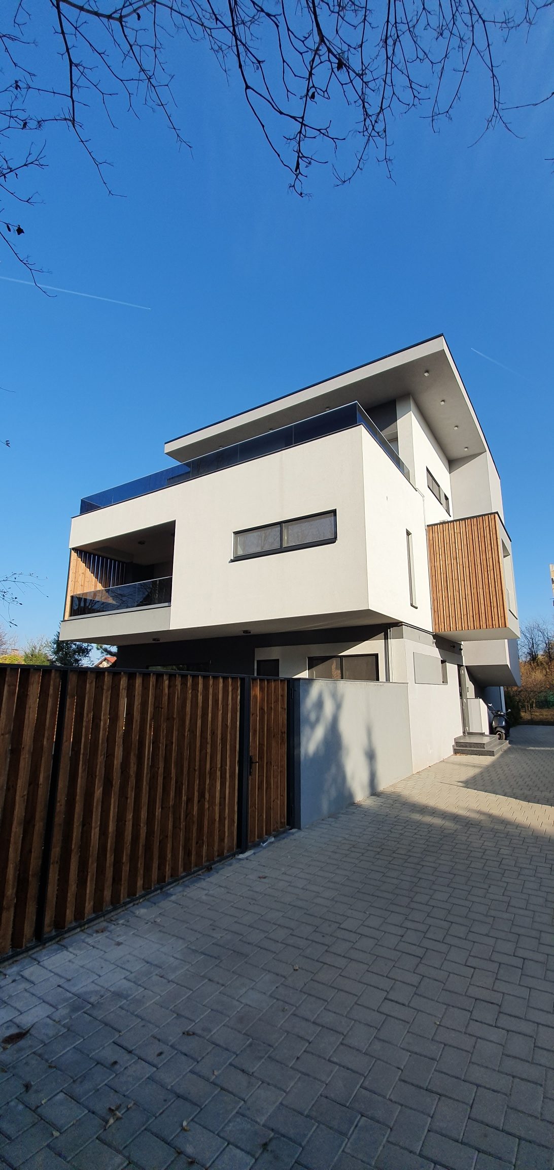 Casa văzută dinspre poartă, cu volumele albe suprapuse, placajul de lemn și aleea pavată