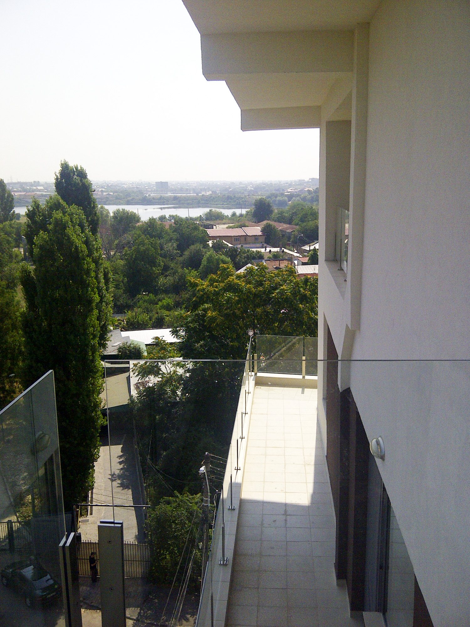 Balconul lung cu parapet de sticlă și priveliștea de la etaj peste case și arbori, spre un lac