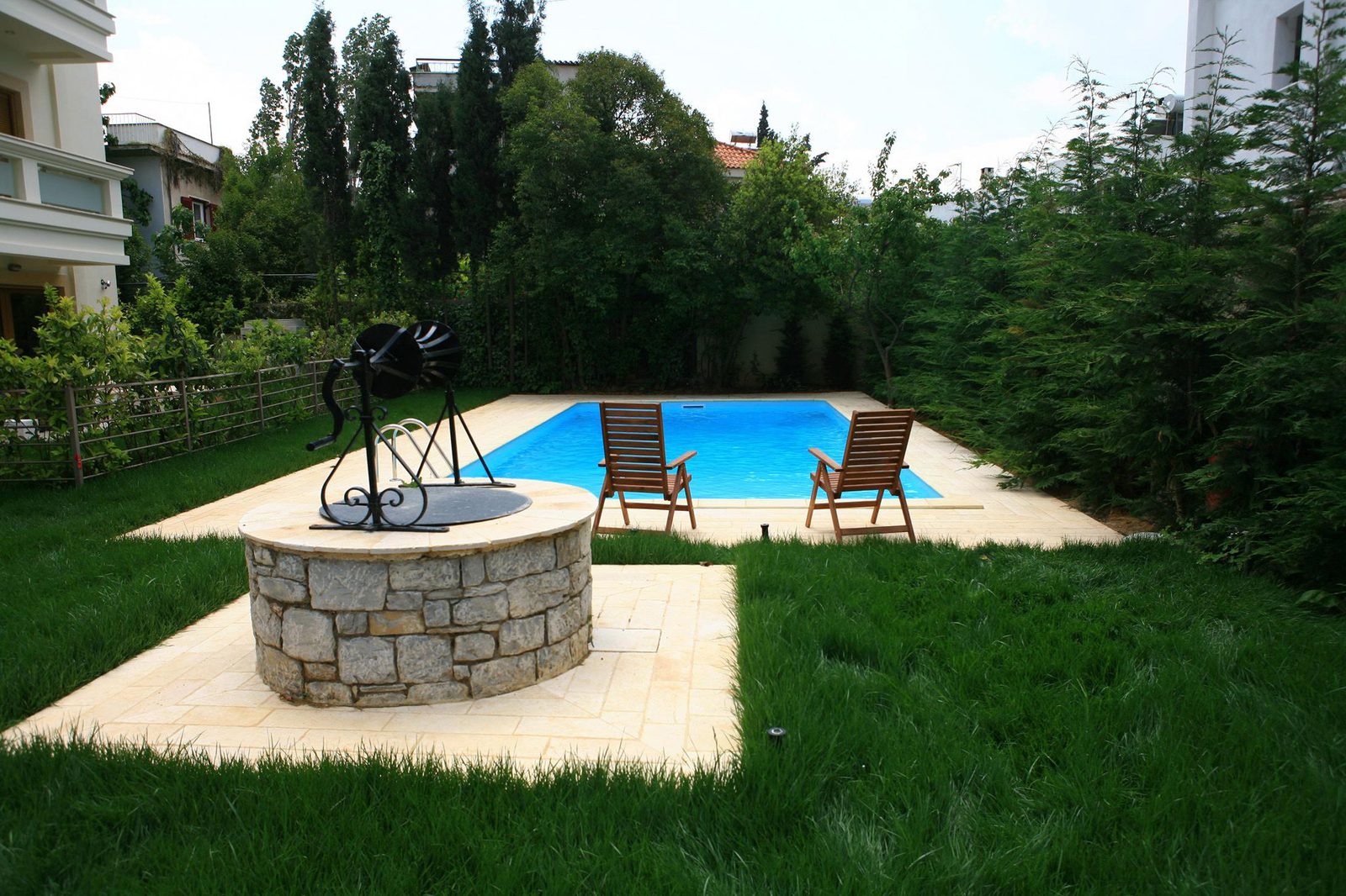 Fântâna de piatră cu roată de fier forjat și piscina din grădină, cu două scaune de lemn